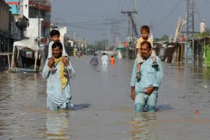 Men carry children on their shoulders and wade along a flooded road, following rains and floods during the monsoon season in Nowshera, Pakistan [File: Fayaz Aziz/Reuters]