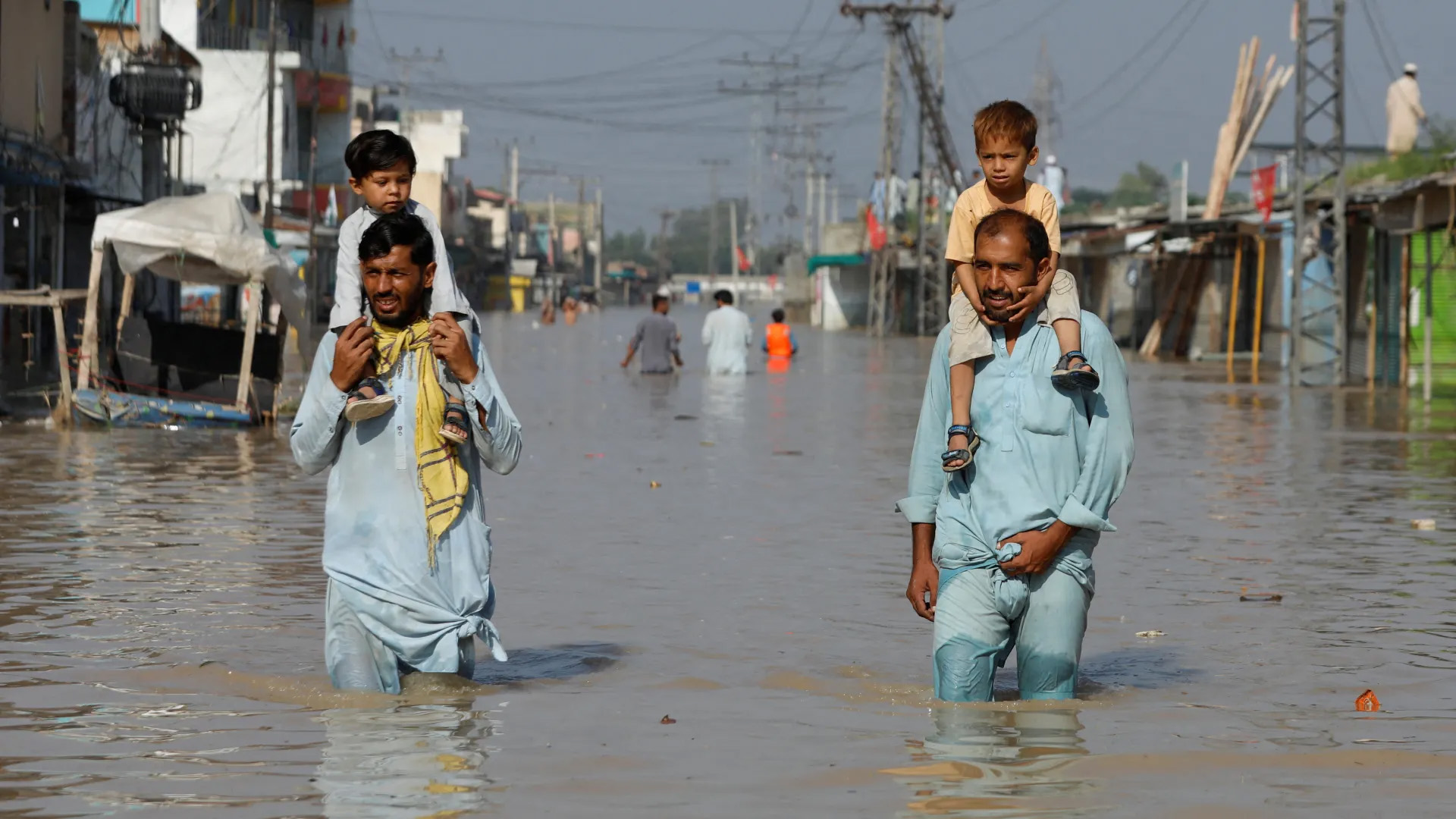 Men carry children on their shoulders and wade along a flooded road, following rains and floods during the monsoon season in Nowshera, Pakistan [File: Fayaz Aziz/Reuters]